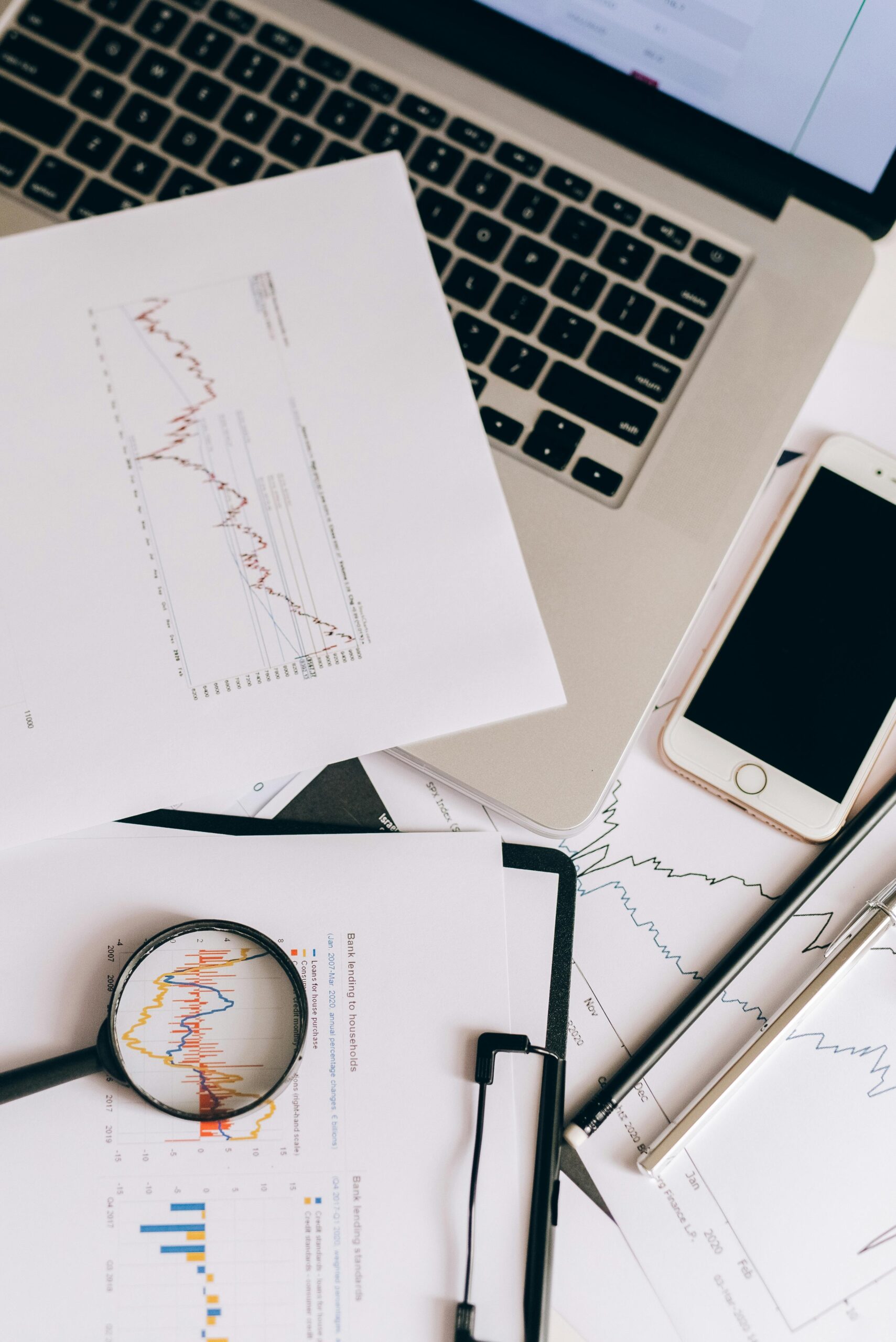 Flat lay of office desk with graphs, laptop, smartphone, and magnifying glass for data analysis.